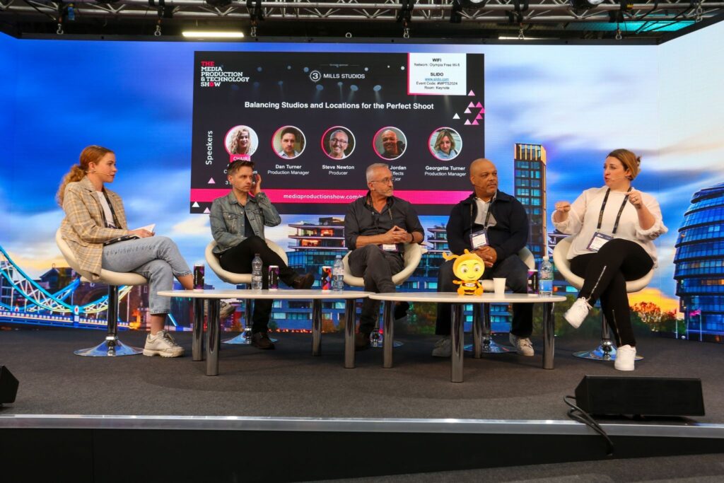 A photo from the panel, 5 people sat in chairs talking on stage.  Behind them on a screen is the panel card, showing their names and photos as well as the panel title 'Balancing Studios and Locations for the Perfect Shoot'.