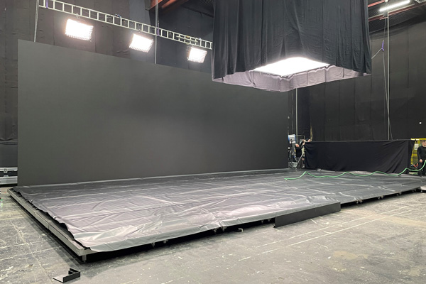 A black sound stage with lighting installed. There is an upright, empty large black screen attached to a floor with black tarp. 