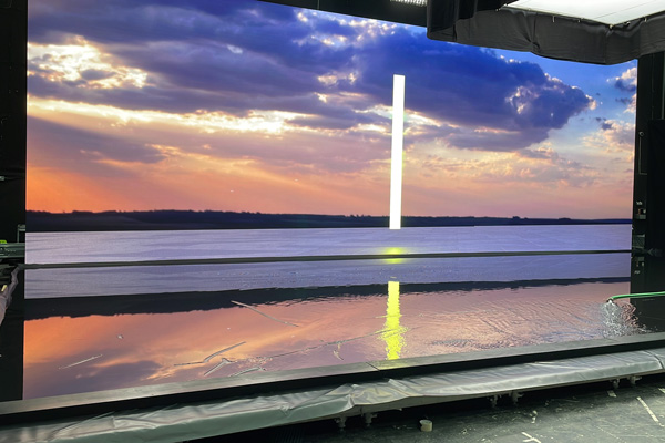 This photo shows the previously black screen now lit up with an image of a orange, blue and purple sunset on a large body of water. The tarp flooring now has water on it which is reflecting the colours from the screen. 