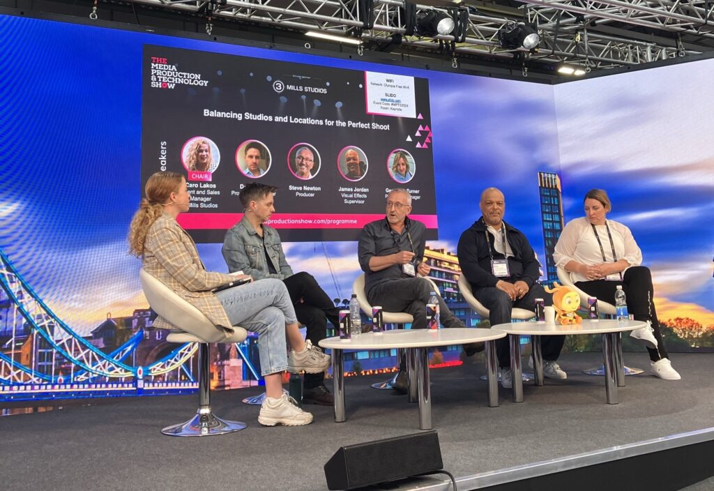 Another shot of the panel on stage in discussion. 5 people are sat in chairs on stage talking. Behind them on a screen is the panel card, showing their names and photos as well as the panel title 'Balancing Studios and Locations for the Perfect Shoot'. 