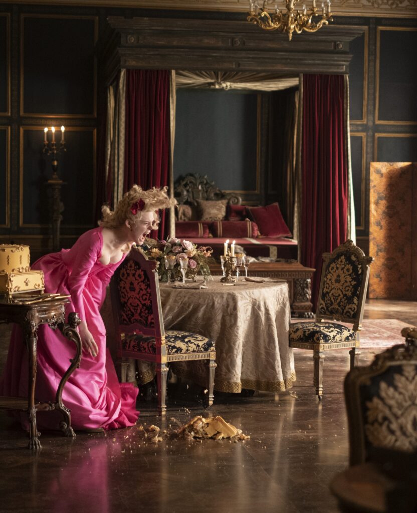 Elle Fanning is screaming and throwing a cake on the floor in a bright pink period costume dress on the set of The Great. Elaborate period furnishing for a bedroom is in the background.